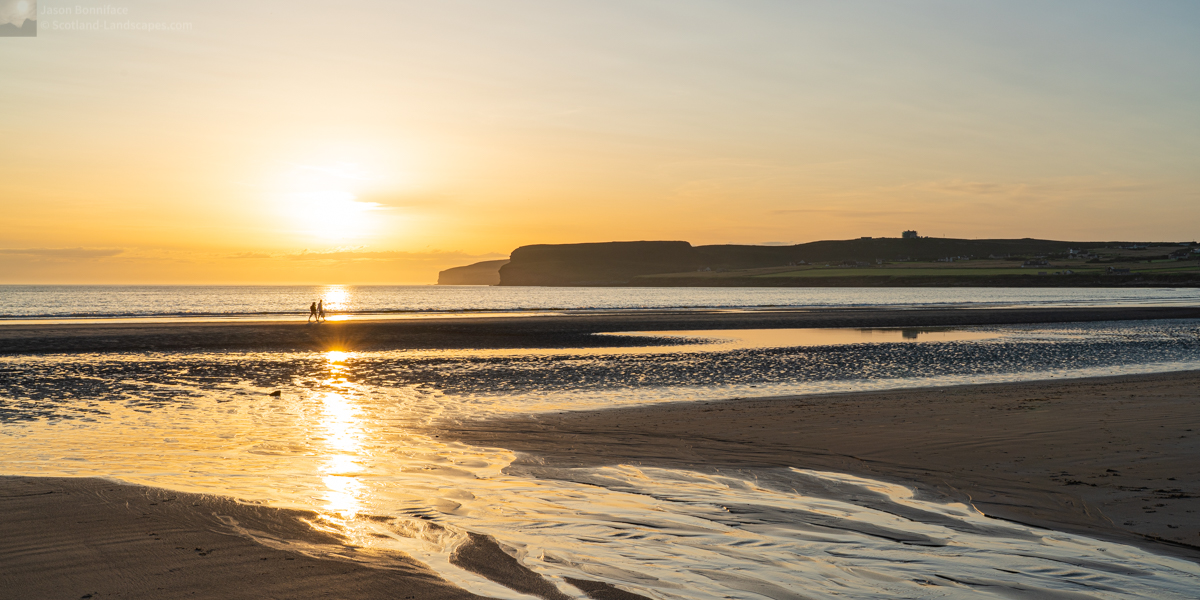 Photo of a couple strolling along the shoreline at Dunnet Beach as the sun descends